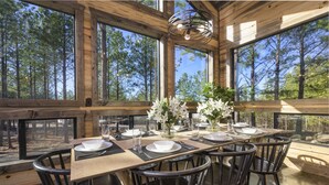 A wooden dining table set for four with chairs, large windows, and a view of trees.