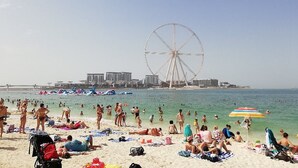 Een strand met mensen die zonnebaden en zwemmen, een reuzenrad en gebouwen op de achtergrond.