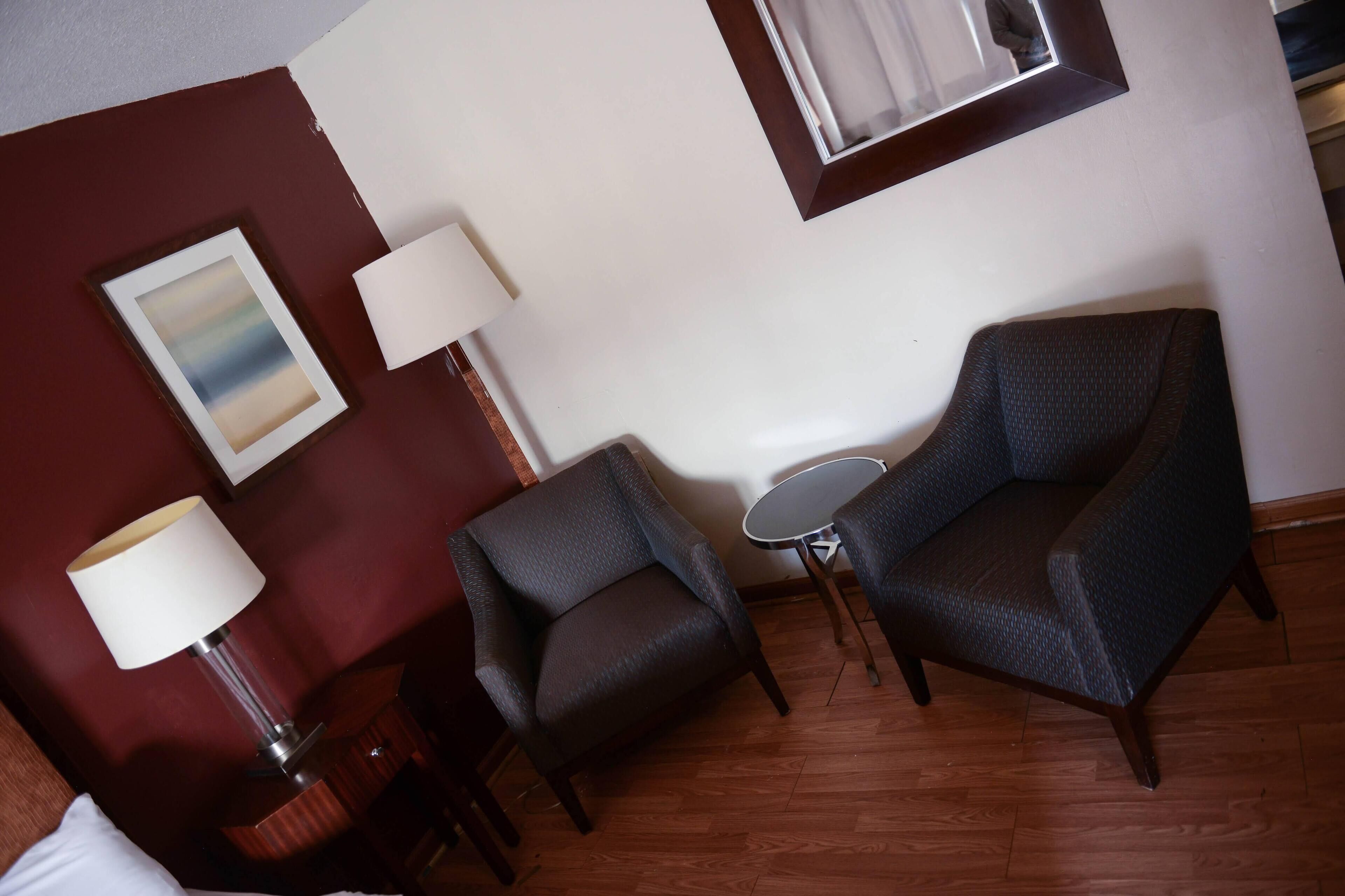 A room with a dark brown wall, a framed artwork, a lamp, two dark armchairs, a small round table, and a window with curtains.