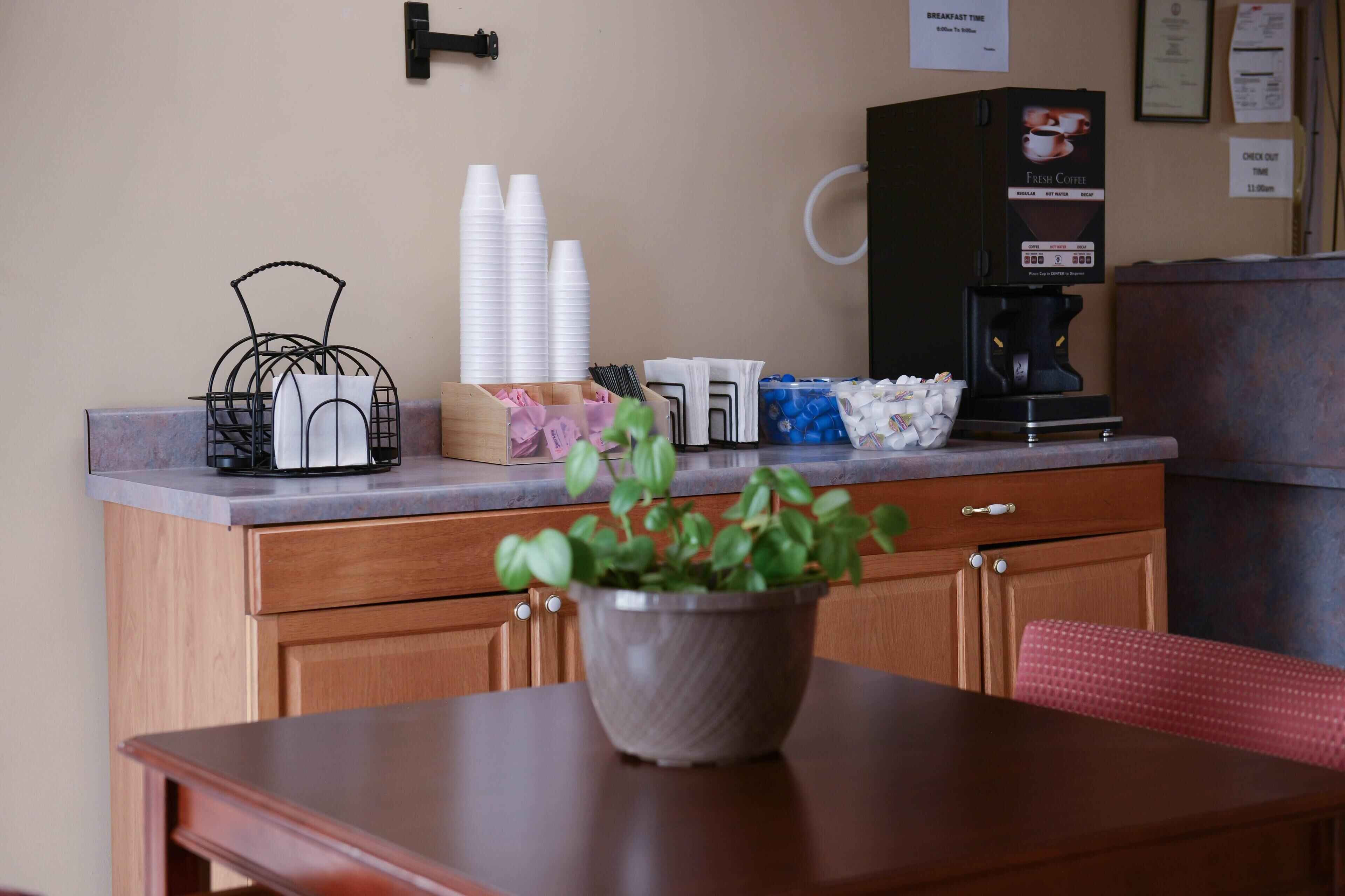 Una kitchenette con una cafetera, vasos de papel apilados y una planta en maceta sobre una mesa de madera.
