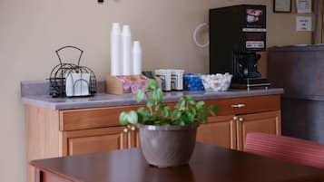 Una kitchenette con una cafetera, vasos de papel apilados y una planta en maceta sobre una mesa de madera.