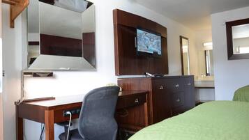 Una camera d'albergo con una scrivania in legno, una sedia, un letto con copripiumino verde, una televisione a parete e uno specchio sopra la scrivania.