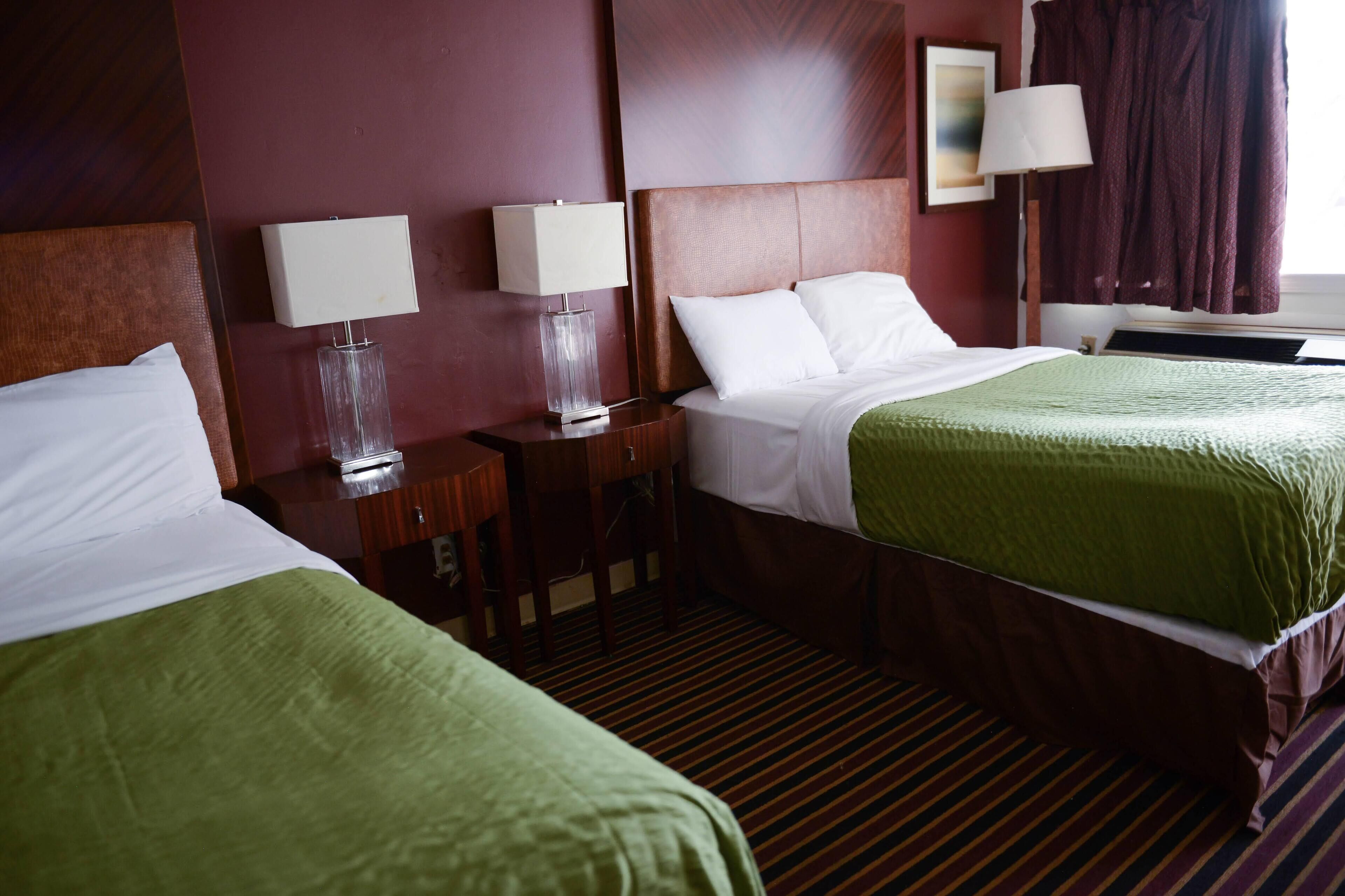 A hotel room with two beds, each with a green bedspread and white linens, a wooden headboard, and a nightstand with a lamp.