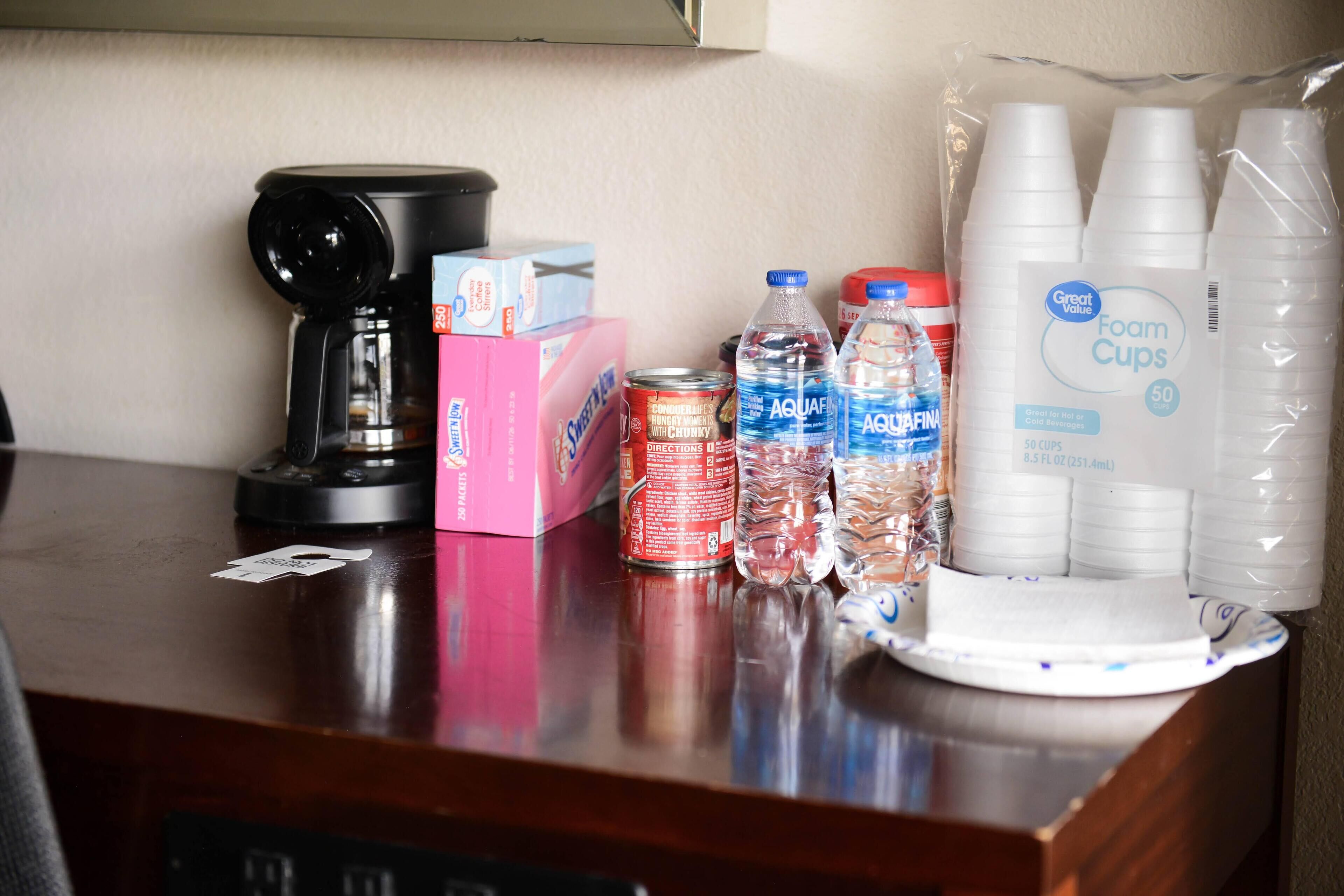 A table with a coffee maker, boxes of tissues, canned drinks, bottled water, and foam cups.