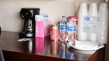 A table with a coffee maker, boxes of tissues, canned drinks, bottled water, and foam cups.