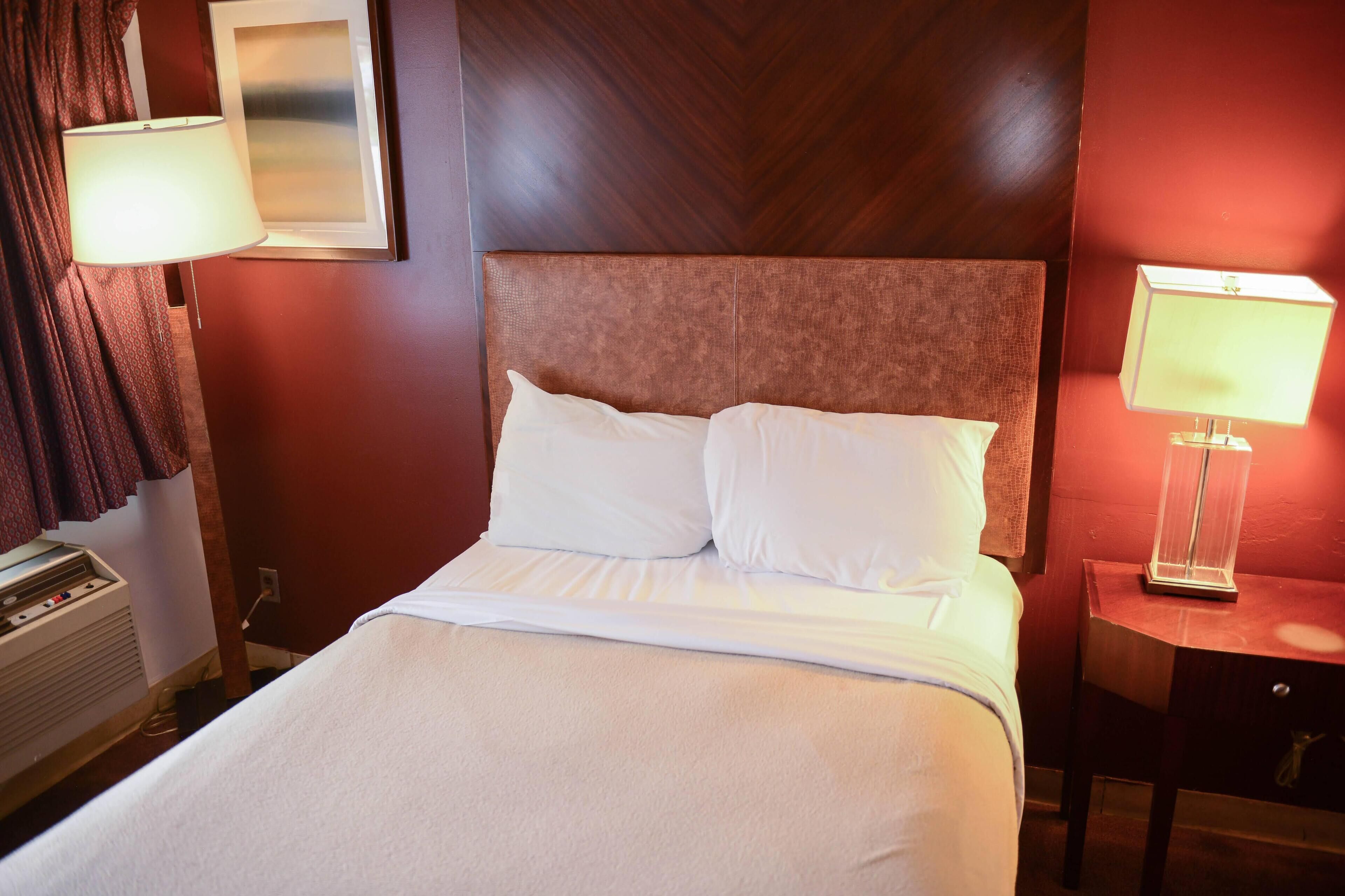 A hotel room with a large bed, two bedside lamps, a wall-mounted air conditioner, and a framed picture on the wall.
