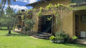 Un edificio de dos pisos con un porche cubierto, un pequeño jardín con plantas en macetas y una palmera al fondo.