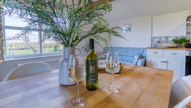 Una mesa de comedor con dos copas de vino y una botella de vino blanco, una planta en maceta, y una vista de un sofá azul y una cocina al fondo.