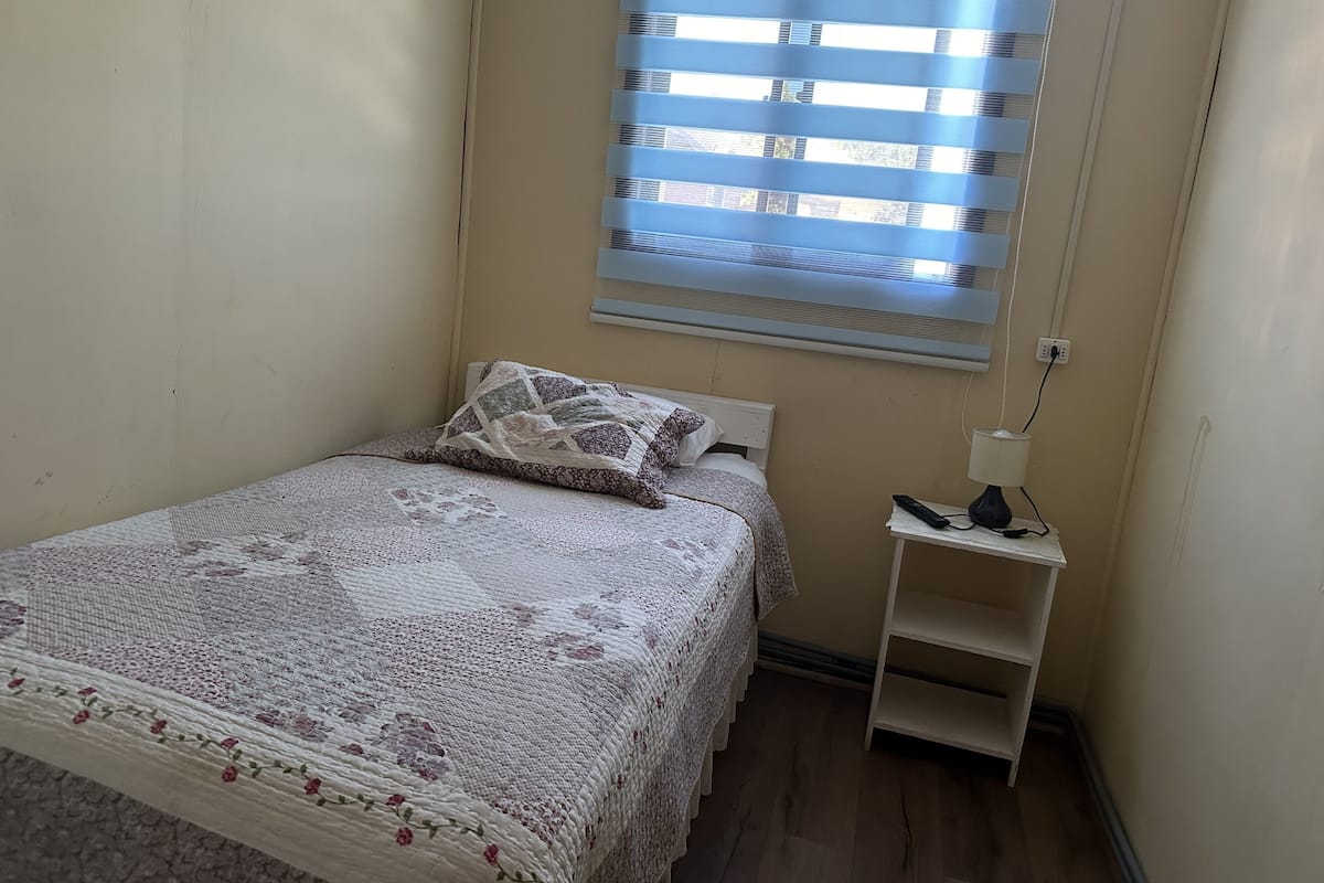 Un dormitorio pequeño y ordenado con una cama individual, una mesita de noche con lámpara y una ventana con persianas.