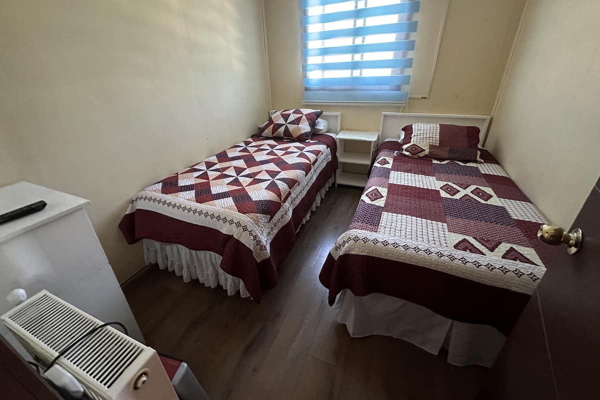 Una habitación pequeña y limpia con dos camas, una ventana con persianas, un radiador y una mesita.