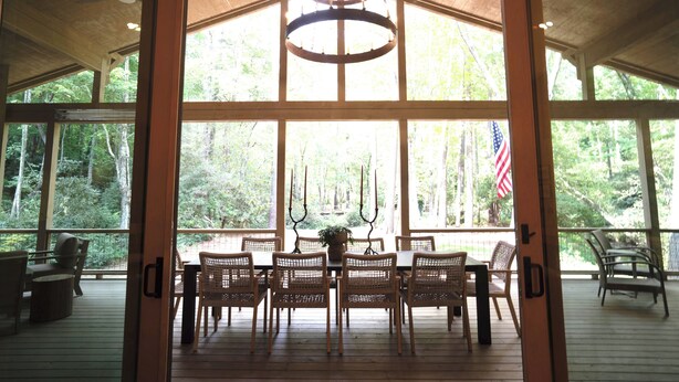 A covered outdoor dining area with wicker chairs and a table, large windows, and an American flag. - Highlands Cabin on Big Creek in Horse Cove. (Highlands)