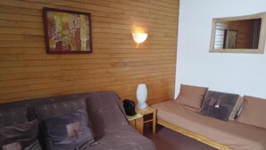 Ett rum med en brun soffa, en mindre divansoffa, ett träbord vid sängen med en lampa och en inramad tavla p å väggen.