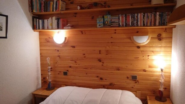 Ett rum med en sänggavel i trä, två sängbord, en säng med ett vitt täcke och en hylla med böcker och dekorativa föremål.