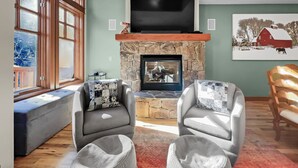 A living room with a stone fireplace, two grey armchairs, a bench, and a large window with wooden frames. - Luxurious Town Home, Short Walk to Lifts, Private Hot Tub 164B (Copper Mountain)