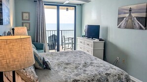 A bedroom with a bed, a desk, a chair, a TV, and a balcony with a view of the ocean. - Beachfront Resort Condo with Pools (Orange Beach)