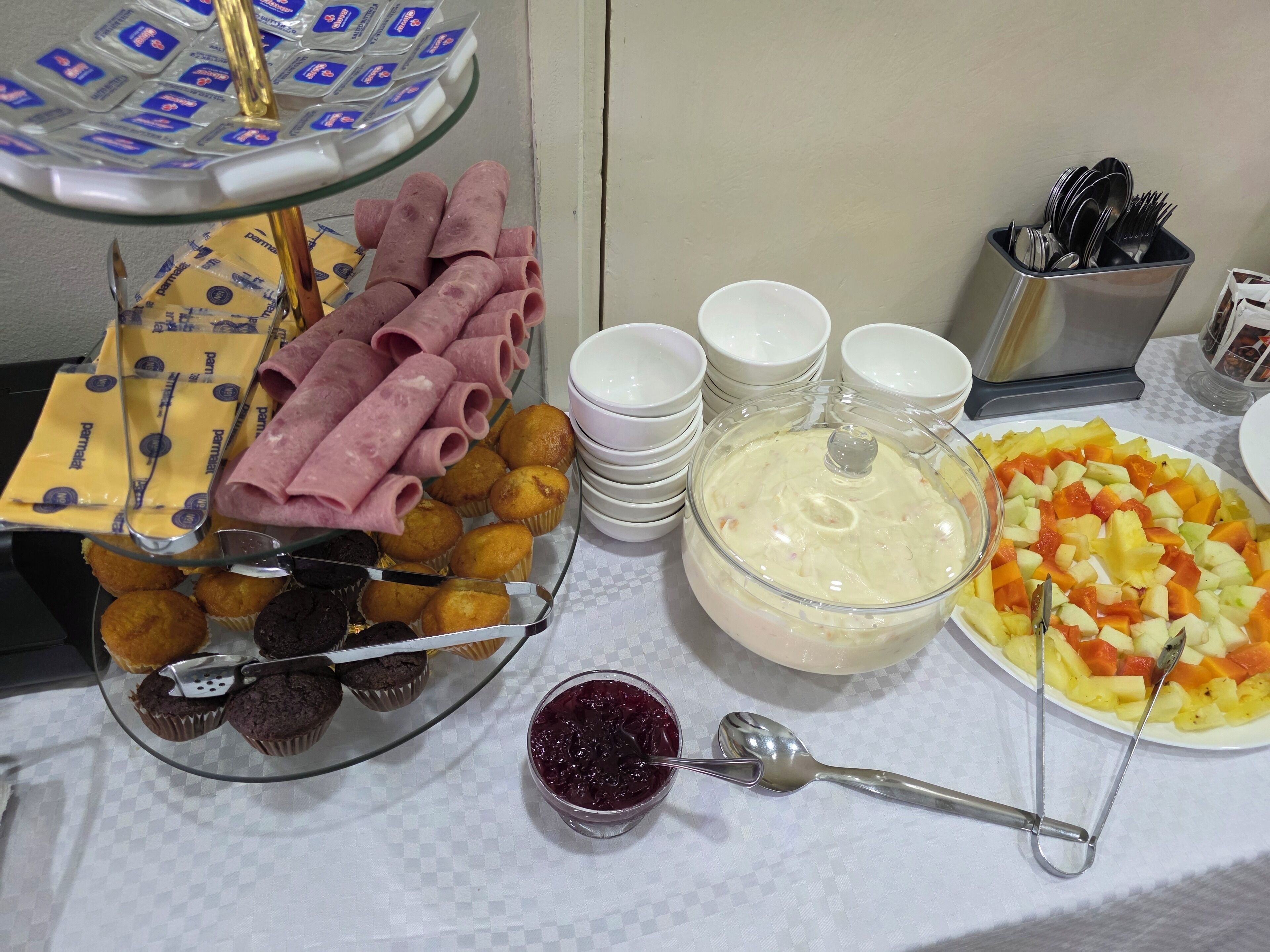 Una mesa de buffet con diversas opciones de comida, incluyendo jamón, muffins y una ensalada de frutas.