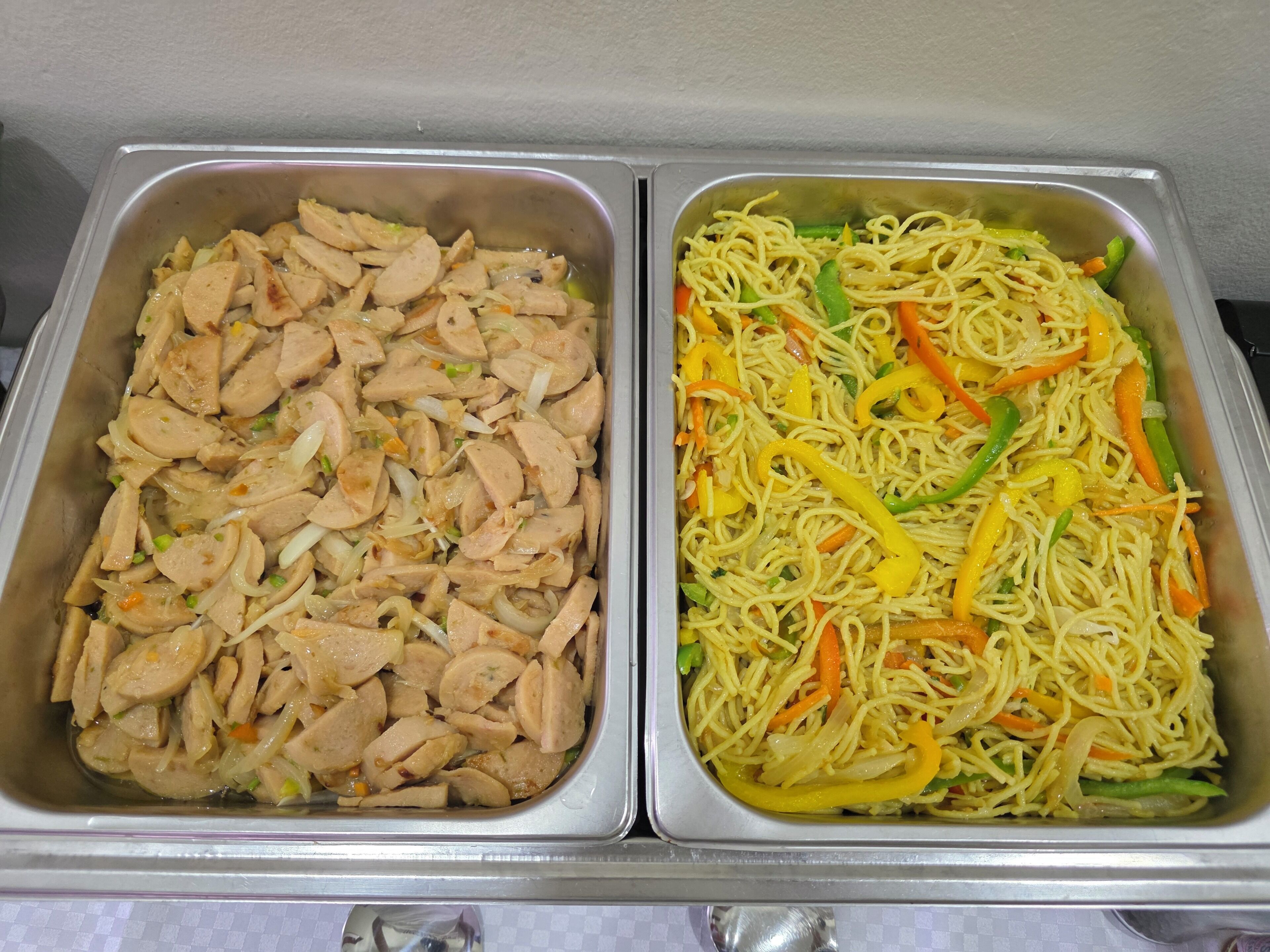 Two stainless steel food service trays, one containing sliced chicken and the other containing stir-fried noodles with vegetables.