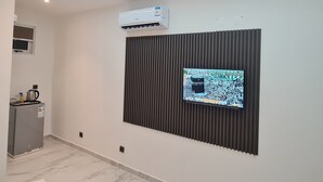 Een moderne hotelkamer met een flatscreen-tv bevestigd aan een geluidsdicht paneel, een ingebouwde airconditioning en een kitchenette met een waterkoker, en een vloer met een marmerpatroon.
