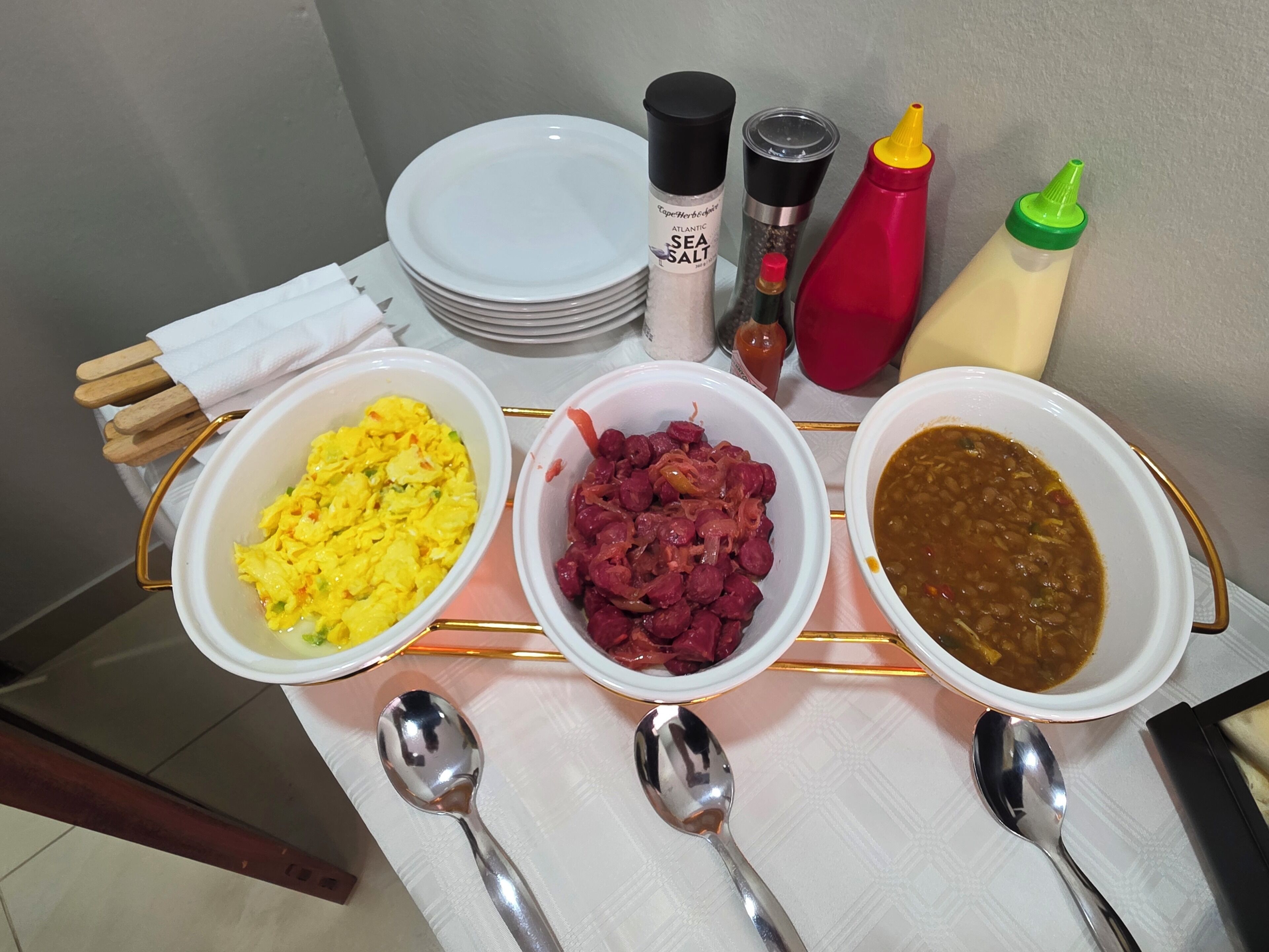 A table with three bowls of food, condiments, and a stack of plates.