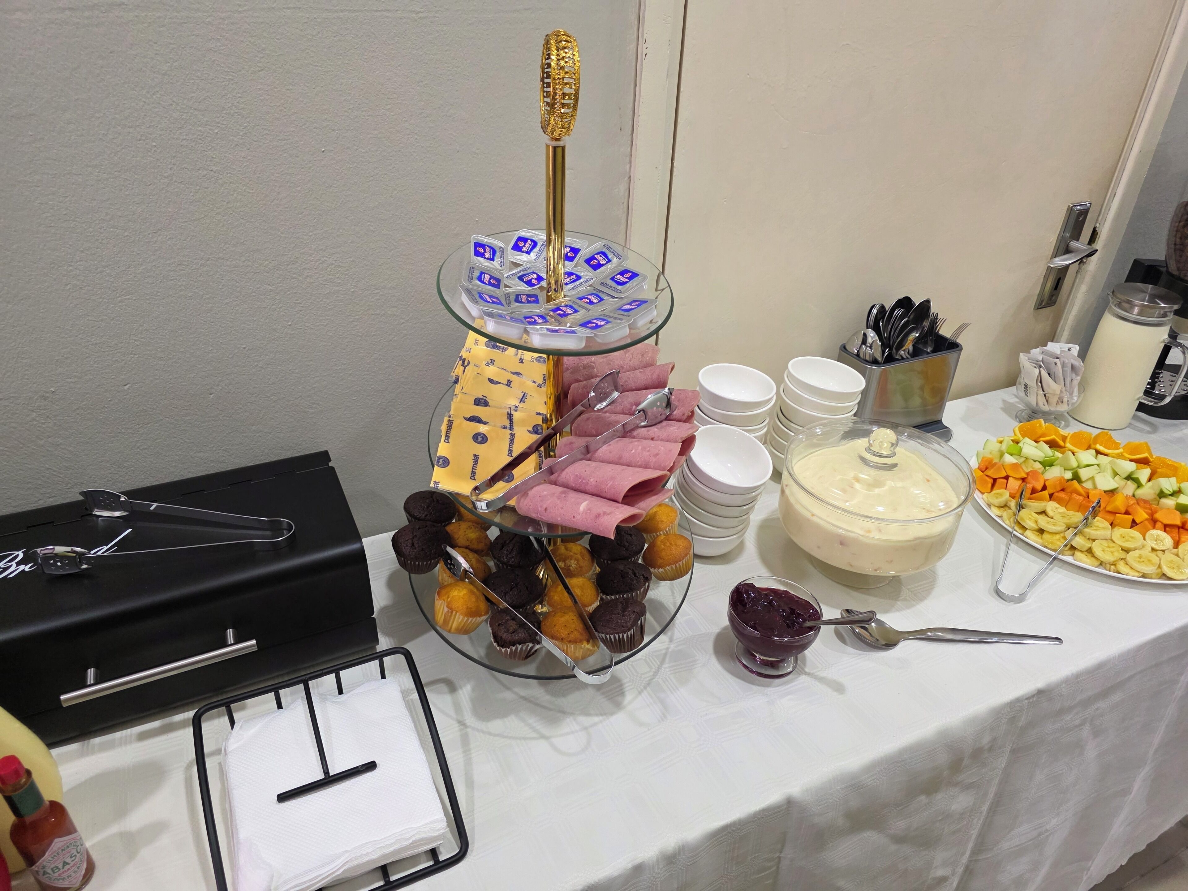 A buffet table with various food items, including a glass stand with pastries, a plate of fruit, and a bowl of dessert.