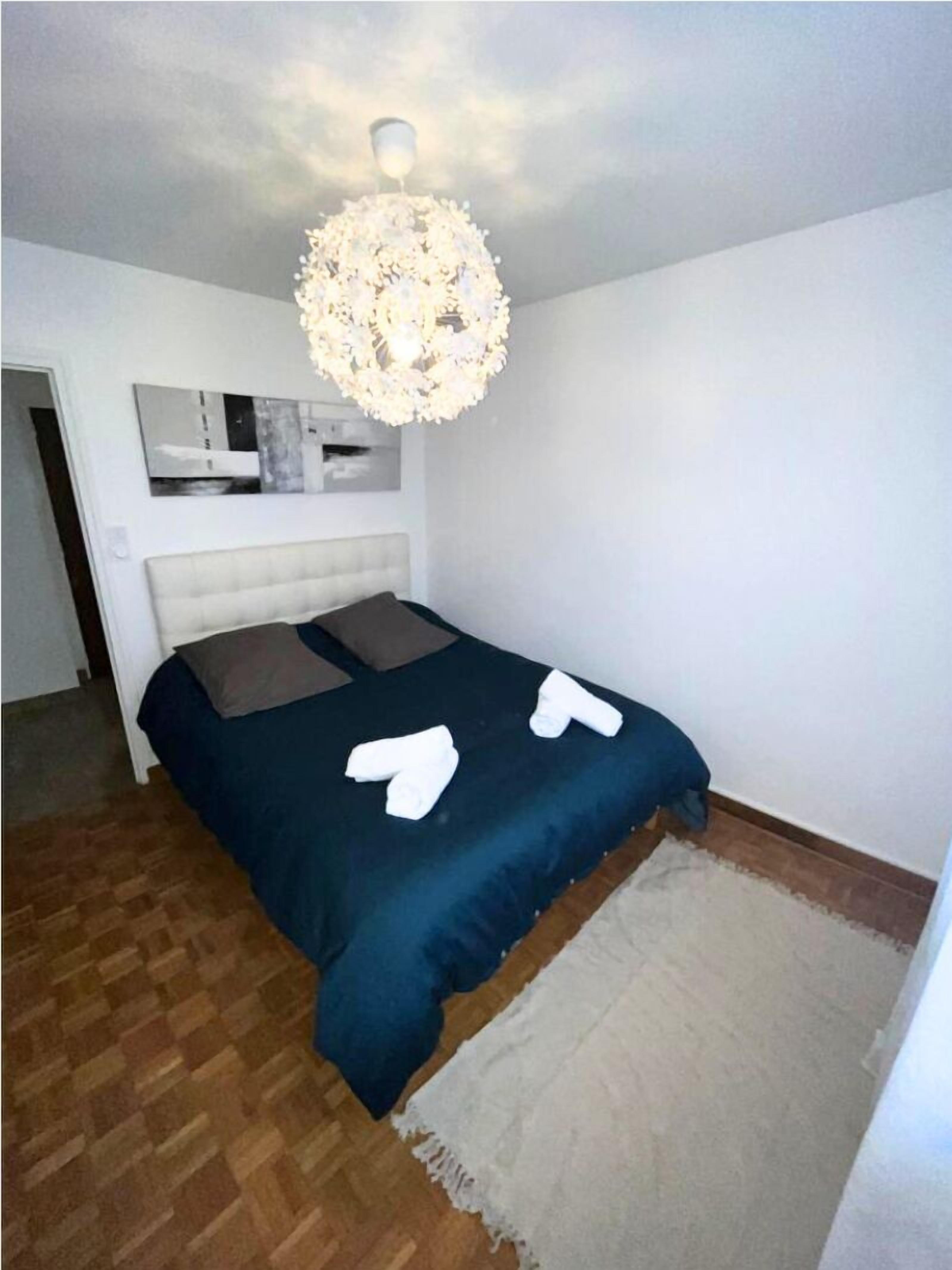 A bedroom with a bed, a white headboard, a large chandelier, and a small wall-mounted mirror.
