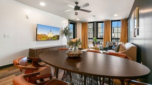 Una habitación de hotel moderna con zona de comedor que incluye una mesa redonda, asientos cómodos, ventilador de techo y ventanas amplias con cortinas.