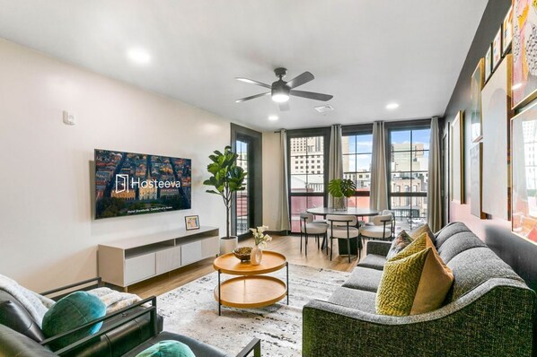 Ein modernes Wohnzimmer mit Flachbildfernseher, Deckenventilator, Essbereich mit Tisch und Stühlen sowie großen Fenstern mit Blick auf die Stadt.