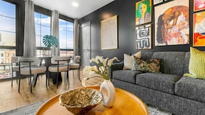 Una sala de estar moderna con un sofá gris, una mesa de comedor con sillas y un ventanal con vistas a la ciudad.