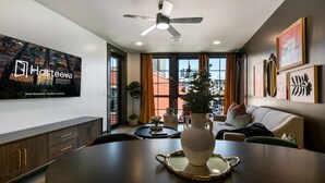 Ein modernes Hotelzimmer mit Essbereich, einem großen Fernseher, der den Namen des Hotels anzeigt, einem Deckenventilator und Blick auf die Stadtlandschaft durch die Fenster.