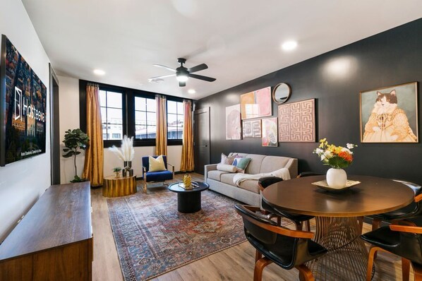 Una sala de estar moderna con sofá, mesa de centro y mesa de comedor. Hay ventanas grandes con cortinas, un ventilador de techo y obras de arte en las paredes.