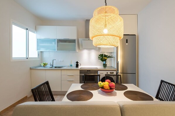 A modern kitchen with white cabinets, a stainless steel refrigerator, a dining table with a fruit bowl, and a large woven pendant light.