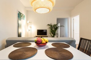 A dining area with a white table, four woven placemats, a bowl of fruit, a grey sofa, a television, and a large plant.