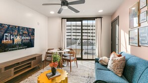 A modern hotel room with a dining area, a sofa, a ceiling fan, and a large window with a view of a parking garage. - Modern 2BR Comfort Gem Condo with 2 Balconies (New Orleans)