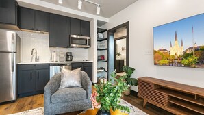 A modern kitchen with dark cabinets, a stainless steel refrigerator, and a microwave. There is a gray sofa with a plant in front of it. A wooden bench is against the wall, and a painting of a building is on the wall. - Modern 2BR Comfort Gem Condo with 2 Balconies (New Orleans)