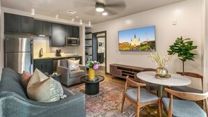 Una sala moderna con un sofá gris, una mesa de centro con un florero, una zona de comedor con una mesa redonda y sillas, una cocina con armarios oscuros y electrodomésticos de acero inoxidable, y un cuadro de un castillo en la pared.