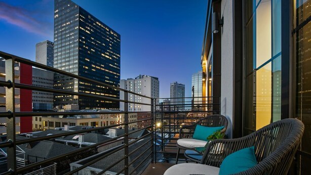Un balcón con sillas de mimbre y una mesa, con vista a un paisaje urbano con edificios altos al anochecer.