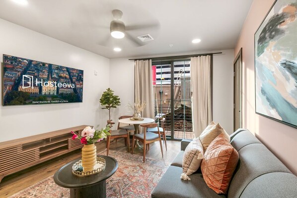 Ein modernes Wohnzimmer mit einer Couch, einem Couchtisch und einem an der Wand montierten Fernseher. Es gibt eine Essecke mit Tisch und Stühlen, sowie ein Balkon mit Blick auf die Stadt.