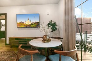 Ein modernes Zimmer mit einem runden Tisch, zwei Stühlen, einer Vase mit weißen Blumen, einem Gemälde einer Kirche und Blick auf eine Brücke durch große Fenster.