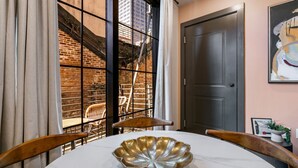 Un comedor con una mesa redonda, sillas, un cuenco decorativo, una ventana con vista a un edificio de ladrillo y una puerta.