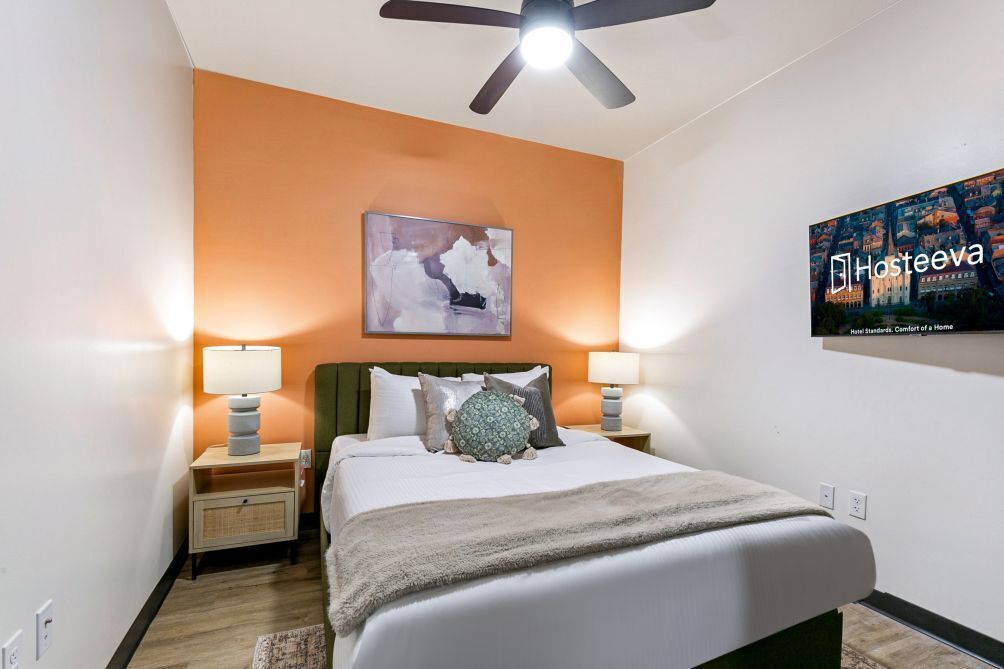 Une chambre d’hôtel comprenant un lit, deux tables de chevet avec des lampes, un ventilateur de plafond, un téléviseur fixé au mur et un tableau.