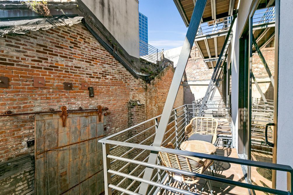 Une ruelle étroite bordée d’un mur de briques, avec un escalier en métal et un petit espace extérieur aménagé avec des chaises et une table.