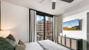 A hotel room with a bed, a ceiling fan, a large window with curtains, a view of buildings, a television on the wall, and a cabinet.