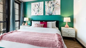 A bedroom with a large bed, teal walls, two abstract horse paintings, and a window with curtains. - Futuristic Luxe: 2BR Urban Gem Condo with Balcony (New Orleans)