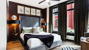 Een moderne hotelkamer met een groot bed, twee bedlampen, een plafondventilator en uitzicht op de stad door grote ramen.