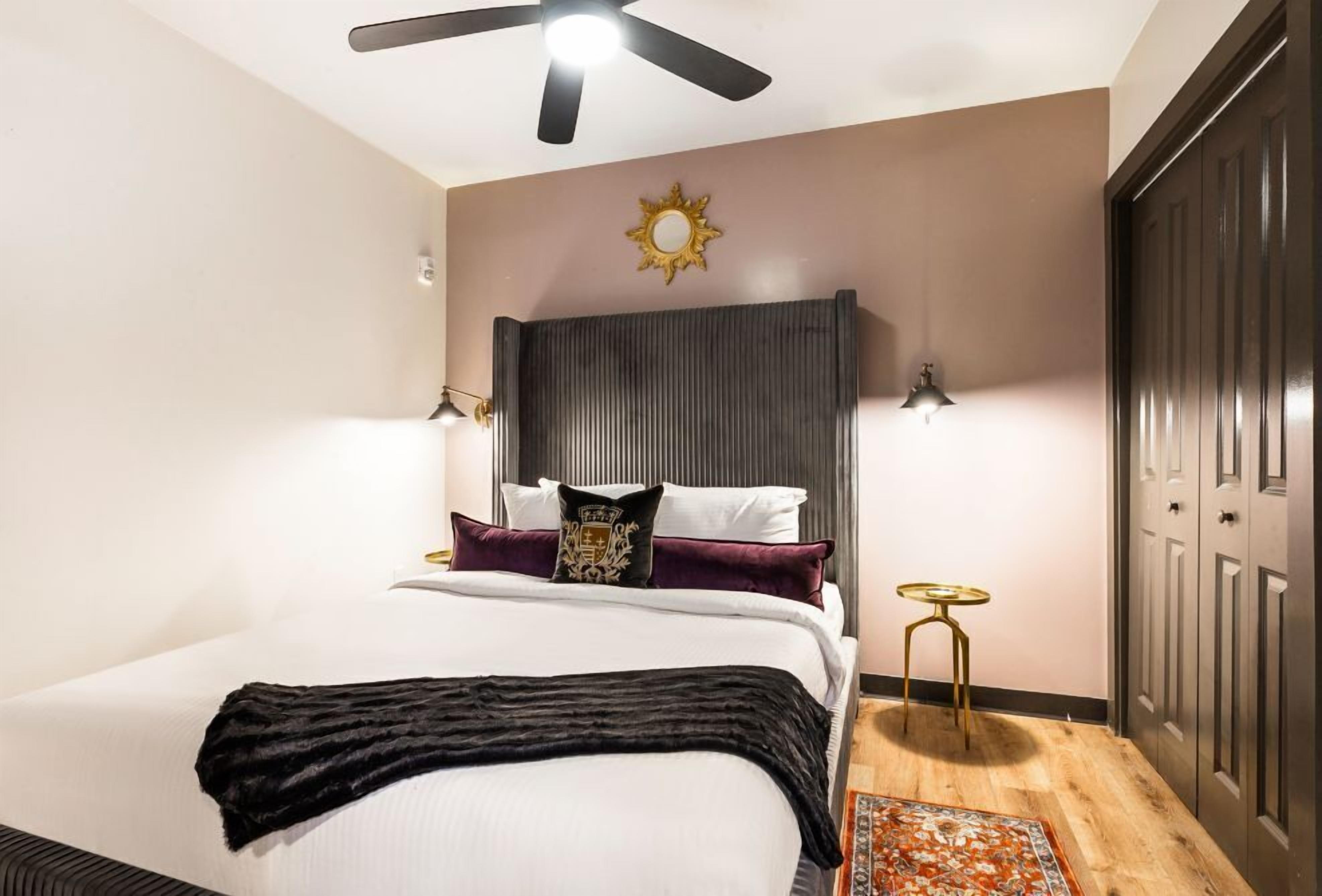 A bedroom with a bed, a ceiling fan, a wall-mounted light, a small table, and a door.