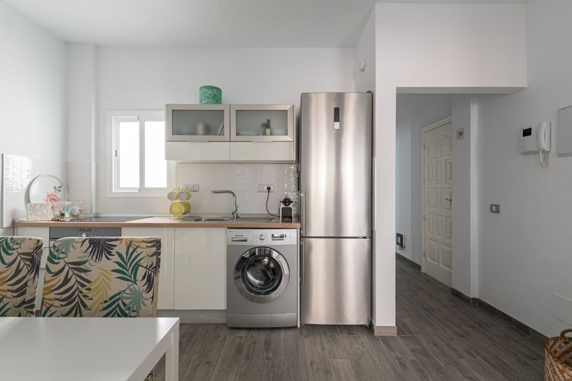 A modern kitchen with a stainless steel refrigerator, a washing machine, and a window above the sink.