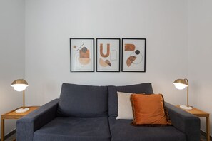 Un salon moderne avec un canapé gris, un seul coussin orange et trois œuvres d’art abstraites accrochées au mur.