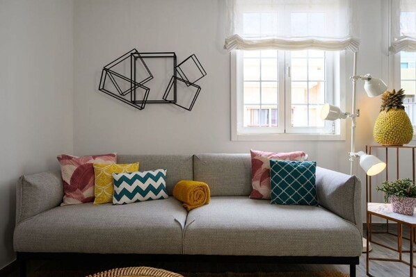 Una sala de estar con un sofá gris, cojines estampados, una obra de arte geométrica en la pared, una ventana con cortinas, una lámpara de pie y una planta en maceta.