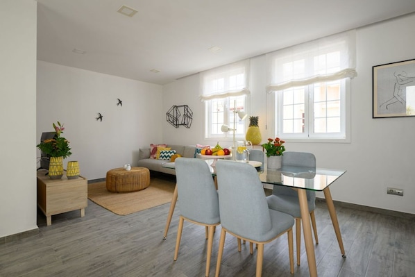 A modern living room with a glass dining table, grey chairs, a sofa, and a small side table with decorative items.