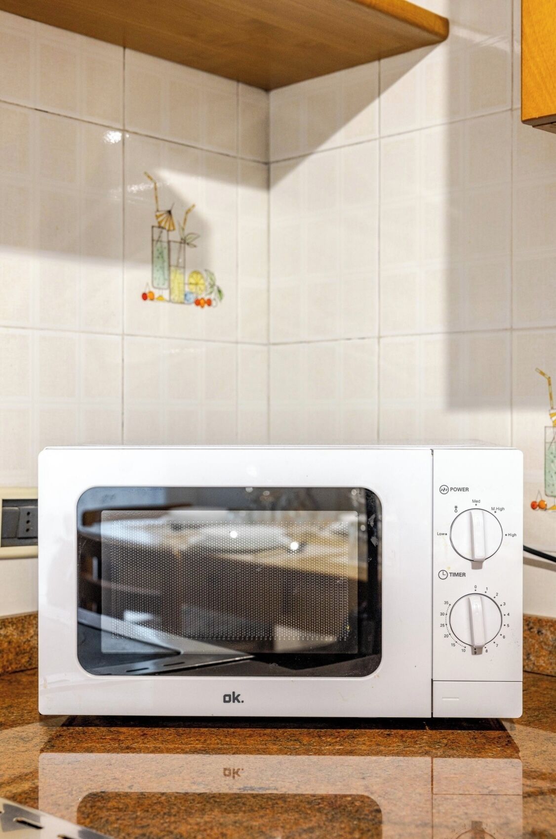 A white microwave oven with a digital display and control knobs for power and timer settings, placed on a granite countertop in a kitchen with white tiles and a wooden shelf.
